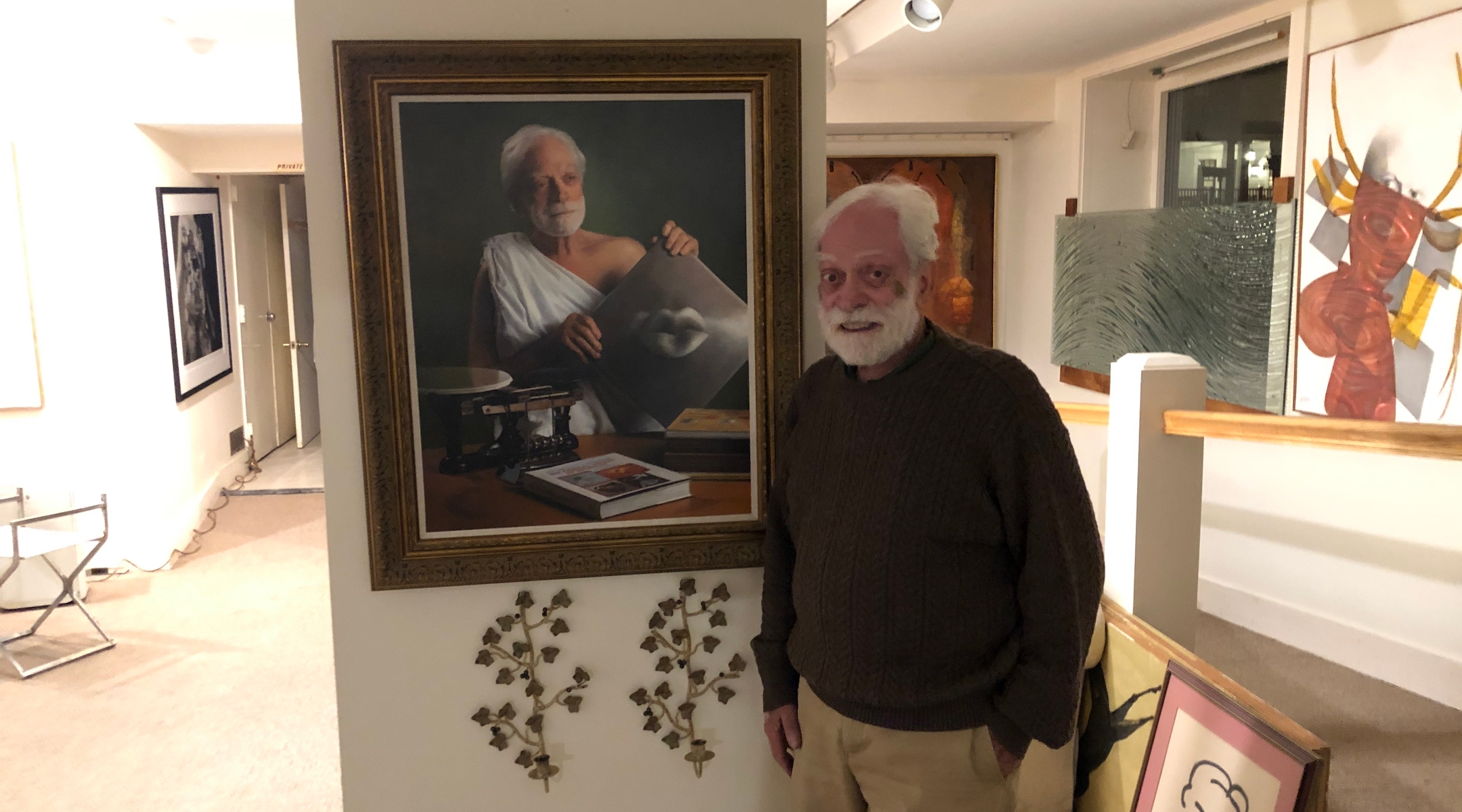 A smiling elderly man poses in front of a Classical-style painting of himself in ancient Greek clothing, posed like a philosopher of old