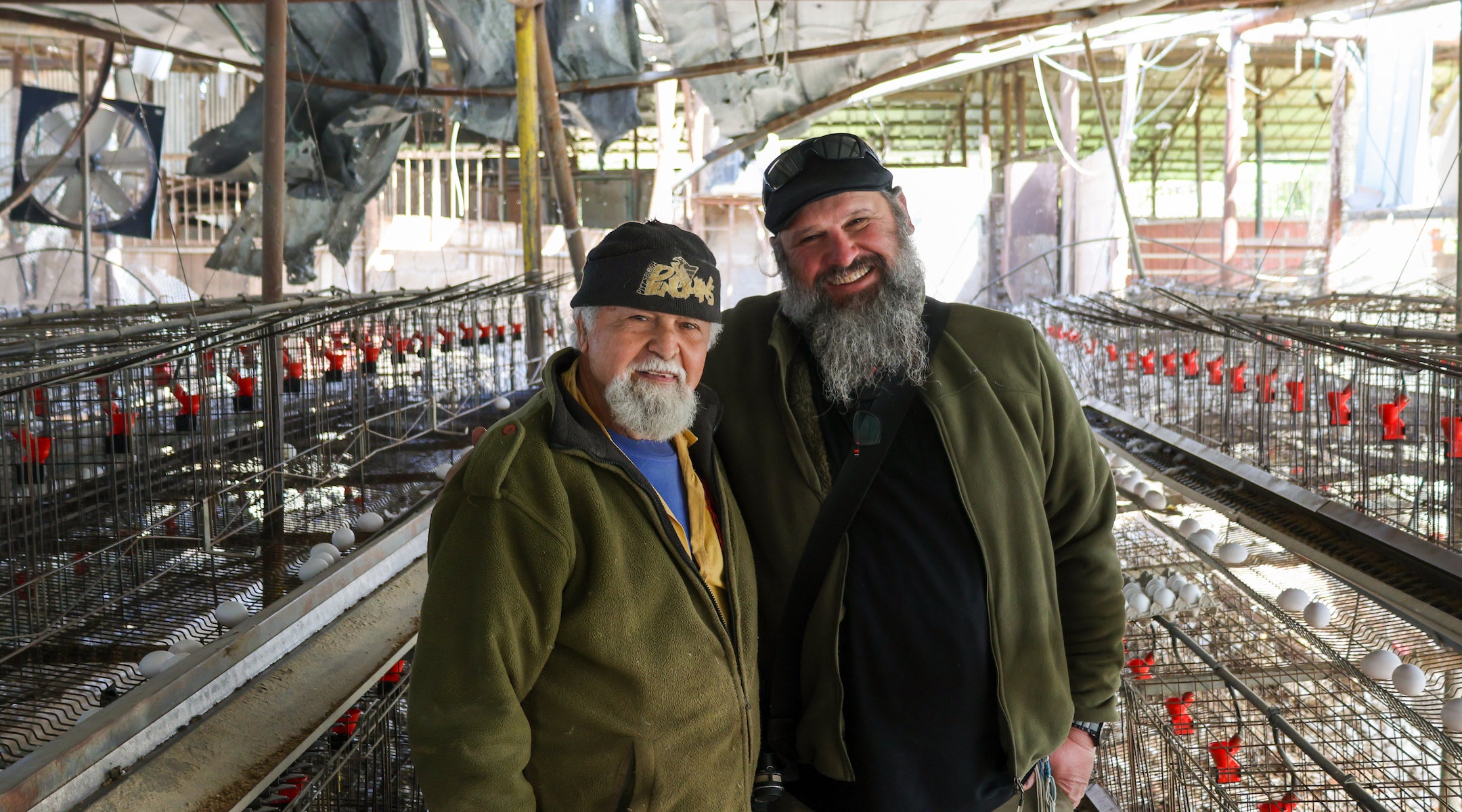 A photo of two men posing surrounded by cages full of eggs.
