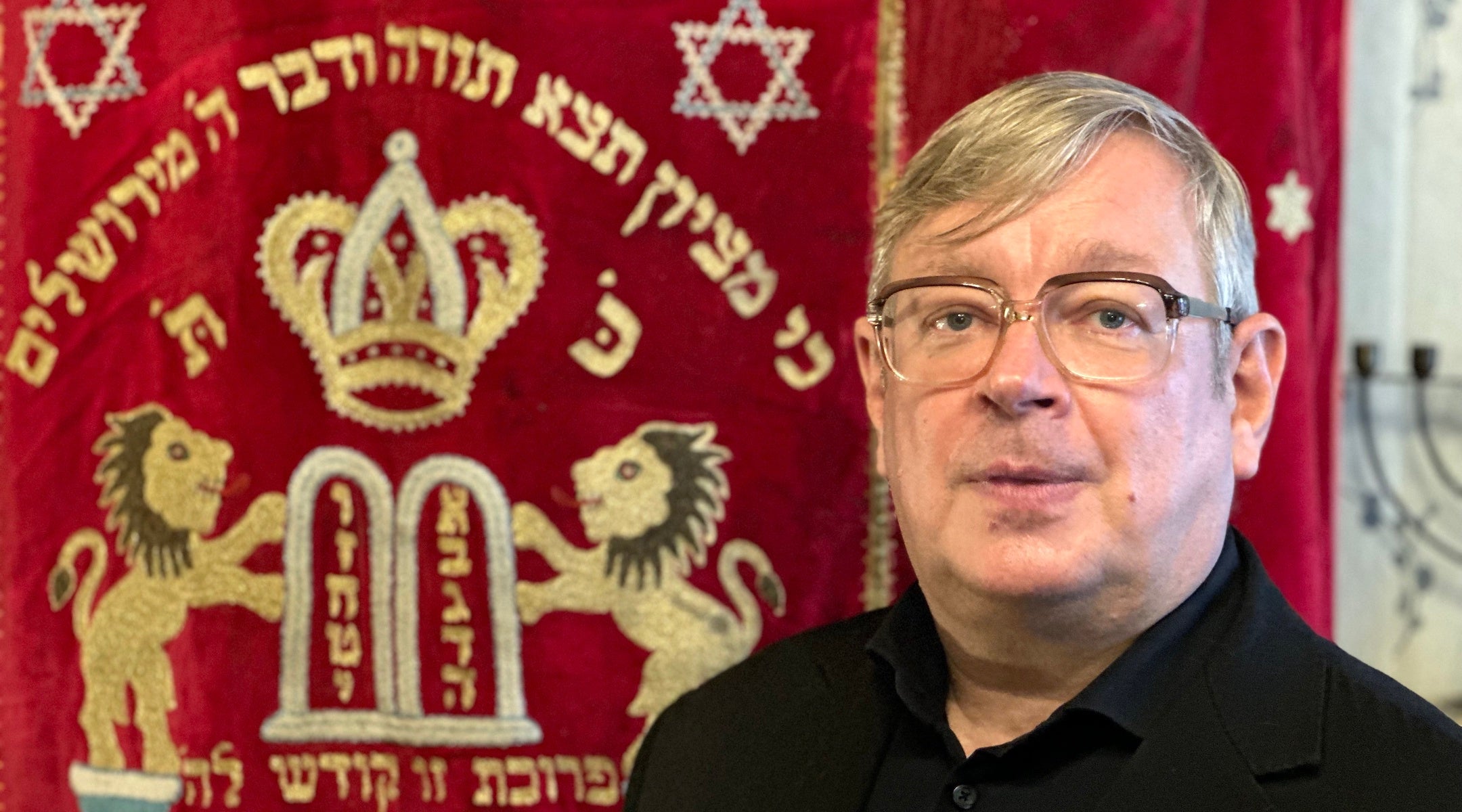 A photo of a man standing next to a tapestry.